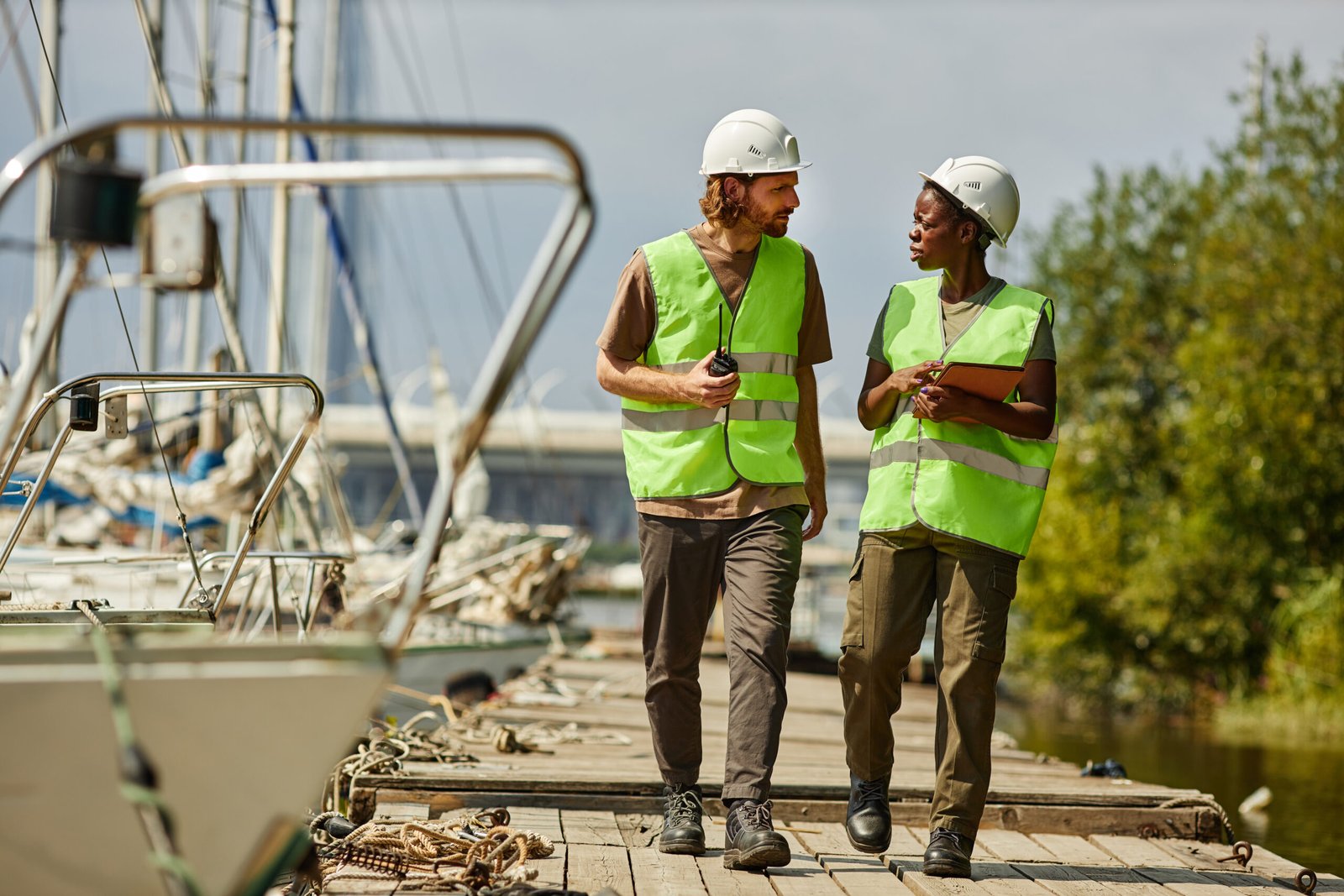 Full length portrait of two young workers wearing hardhats while walking towards camera on pier, copy space