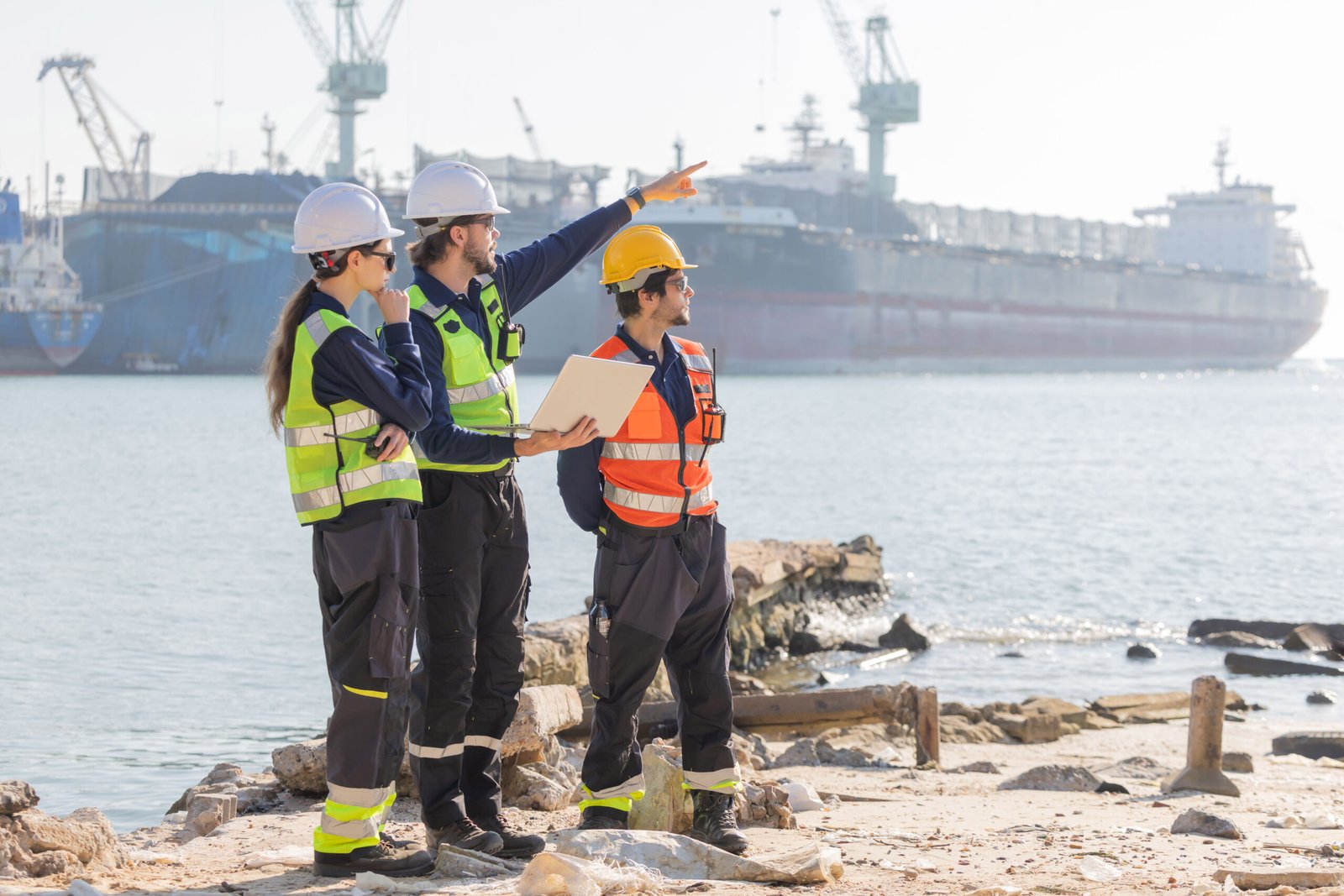 Workers discuss plans at a port with a cargo ship in the background during a sunny day near the water