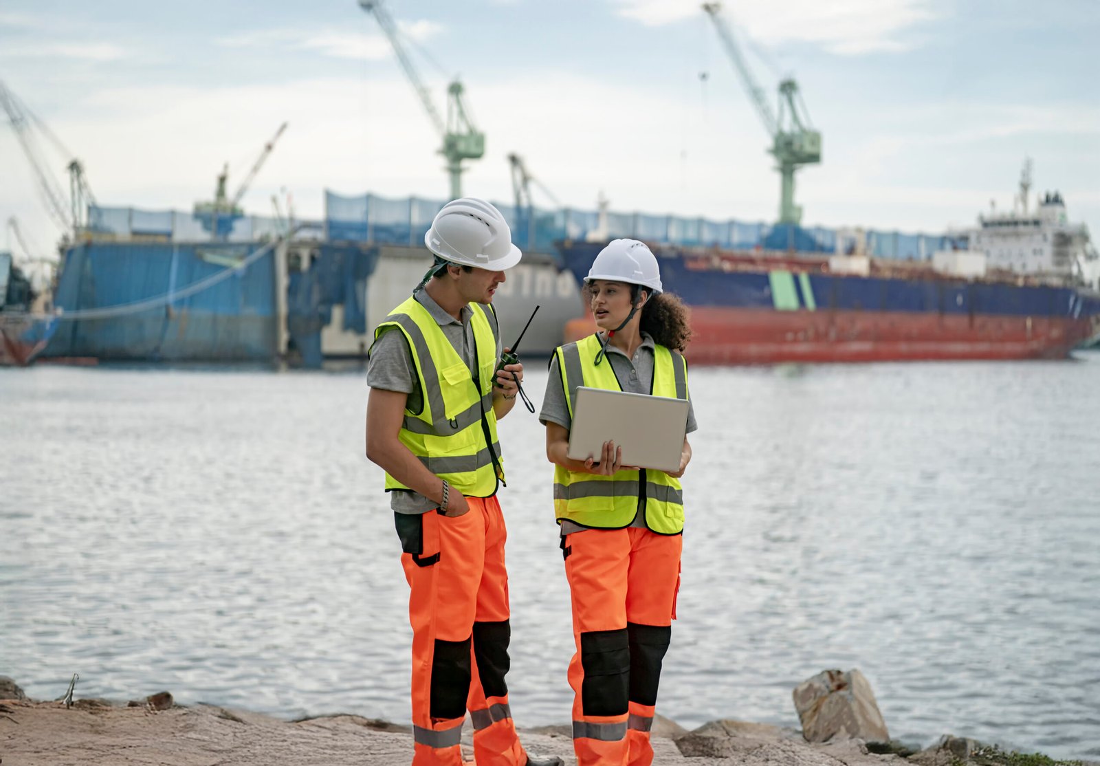 Two construction workers dressed in safety attire talk while using a radio and laptop beside a harbor filled with cargo ships during late afternoon.